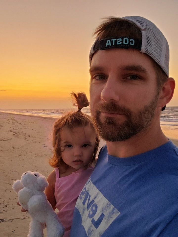 Troy holding his daughter on the beach at sunset